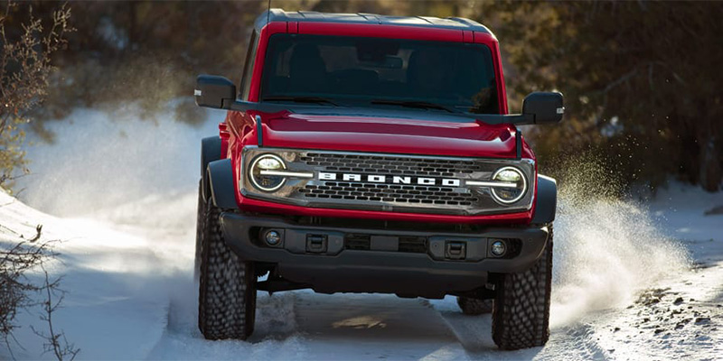 Red Ford Bronco driving through snow-covered terrain, kicking up snow behind.