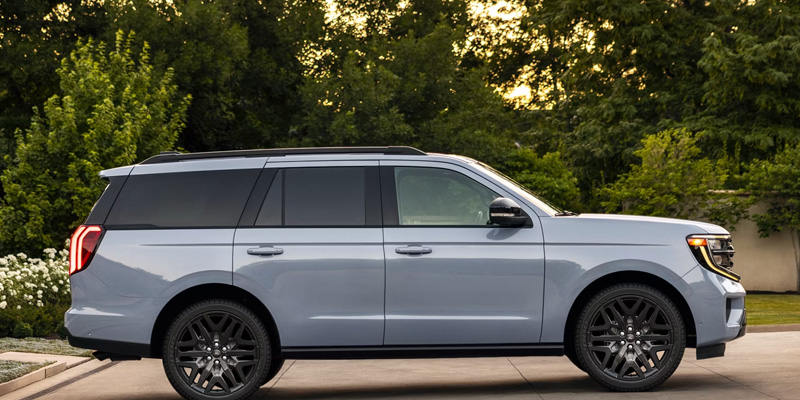 Side view of a silver full-size SUV parked on a driveway with trees and bushes in the background