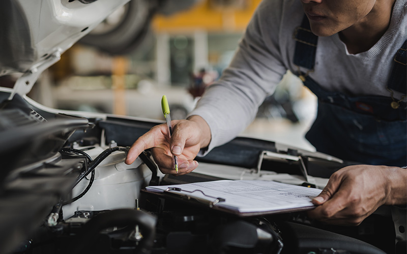 Auto technician inspecting a vehicle engine while holding a clipboard and making notes in a service garage