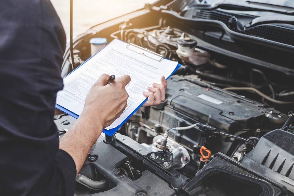 Automotive technician writing on a clipboard while inspecting a car engine bay during a vehicle diagnostic check in a repair shop.