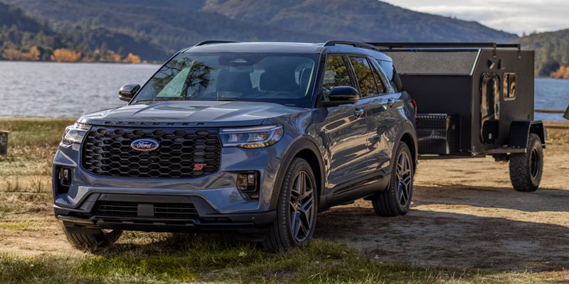 Front three-quarter view of a gray Ford SUV parked near a lake with a camper trailer attached, showcasing outdoor towing capability in a scenic mountain setting.