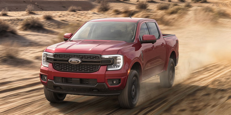 Front three-quarter view of a red Ford Ranger truck speeding through a desert landscape, kicking up sand and dust to emphasize off-road performance.